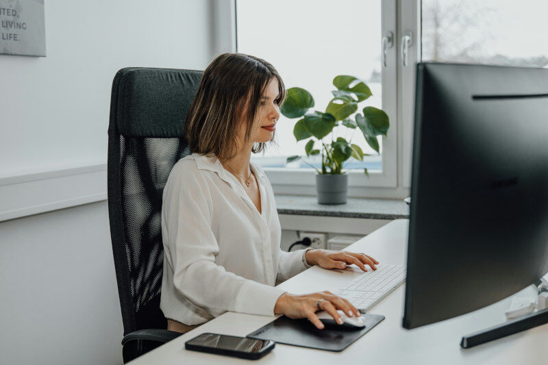 Eine brünette Frau sitzt im Büro an ihrem Arbeitsplatz vor einem großen Bildschirm und beschäftigt sich mit der neuesten Software.