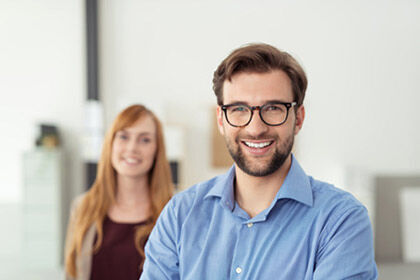 ein Mann im Vordergrund und eine Frau im Hintergrund lächeln